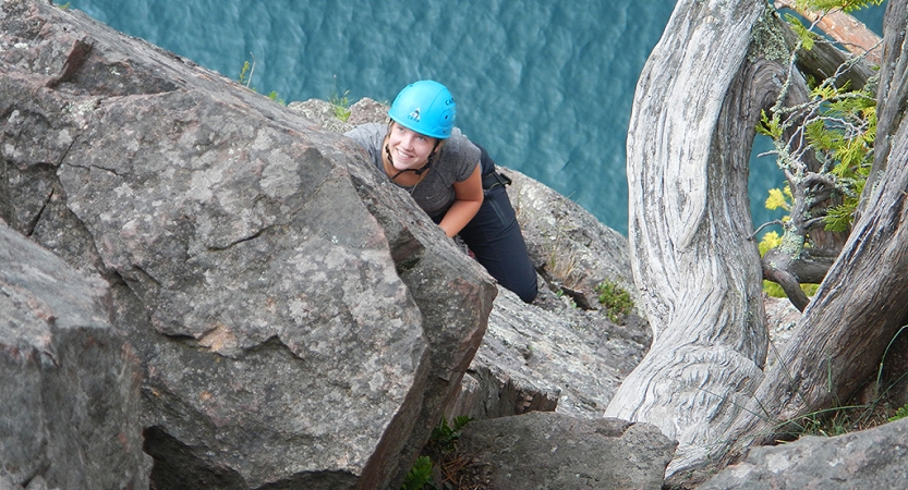 A person wearing safety gear is secured by ropes as they look up at the camera and smile while rock climbing. They are high above a blue body of water. 